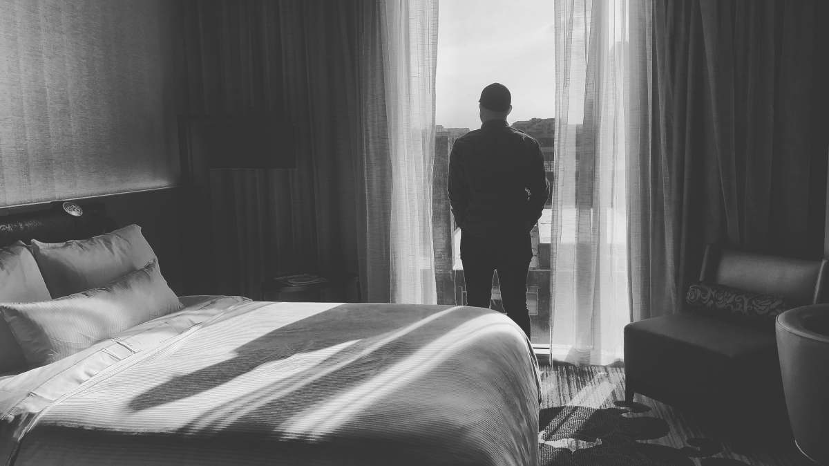 Explore a serene moment captured in a contemporary hotel room, where a thoughtful man stands before a beautifully draped curtain, gazing out the window. This striking scene highlights modern interior design elements, making it perfect for lifestyle blogs, travel magazines, or real estate visuals. The elegant bedding and refined dcor create an inviting atmosphere, ideal for discussions on relaxation and personal space.