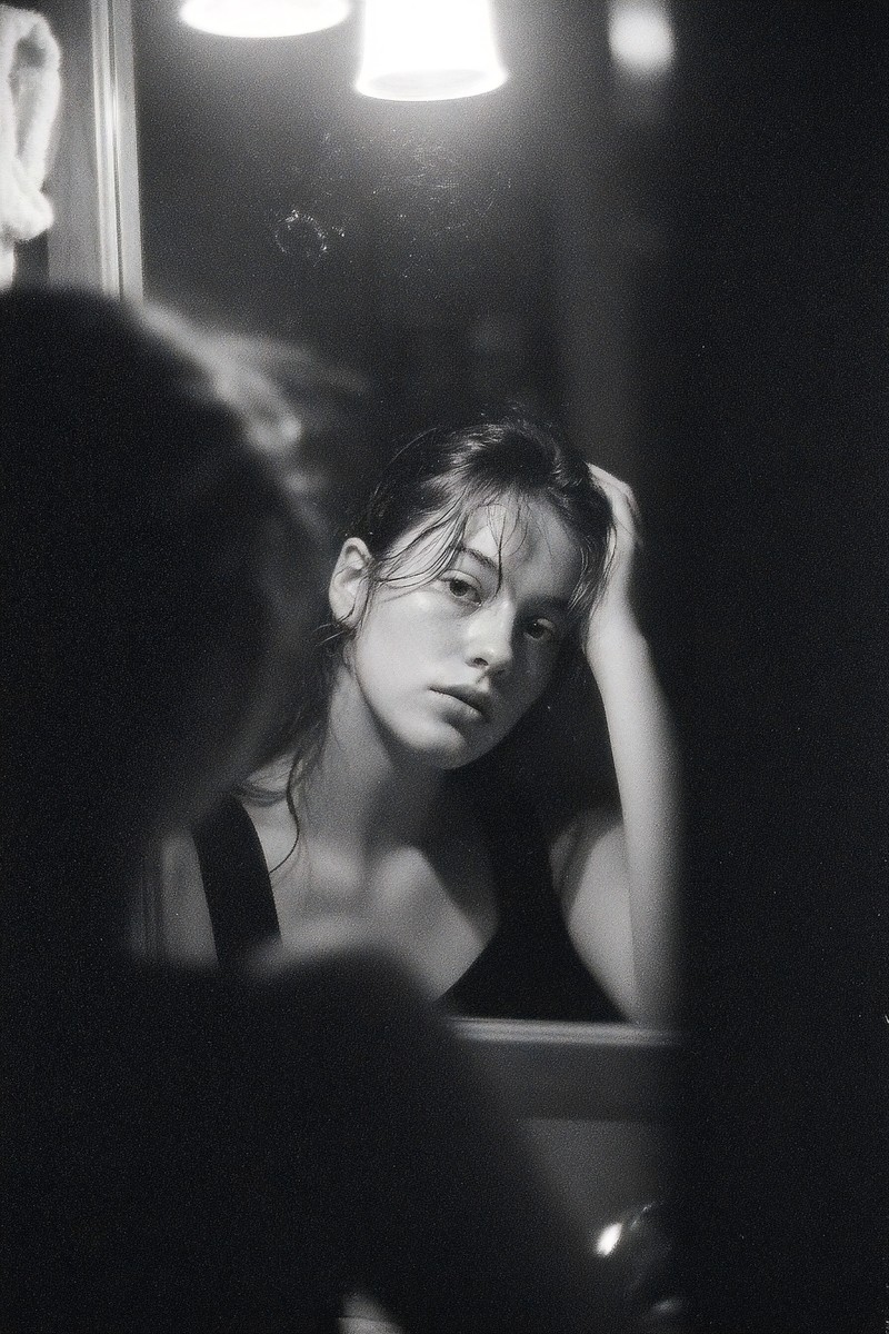 In a dimly lit room, a woman with disheveled hair gazes into a mirror, lost in thought. Her reflection reveals the weight of overthinking, evoking emotions of introspection and self-discovery. This image captures a moment of vulnerability, making it ideal for projects focused on mental health, self-reflection, or personal empowerment, resonating with audiences seeking authenticity.