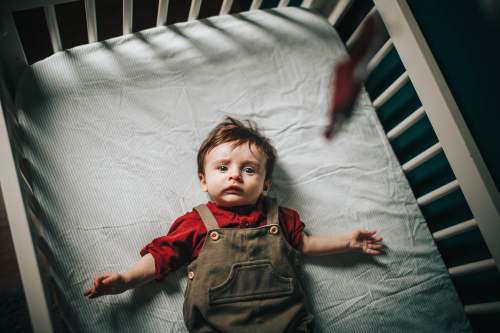 grey girl in red button up shirt and black denim vest lying on white bed baby in crib